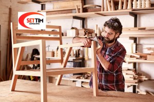 Serious furniture designer carefully sanding a chair frame that he is busy manufacturing in his woodwork studio, with shelves of wooden items behind him
